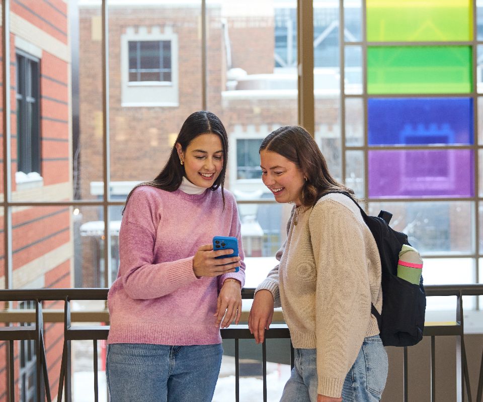Two college students engage with a smartphone while smiling, standing in a brightly lit corridor with colorful windows in the background.
