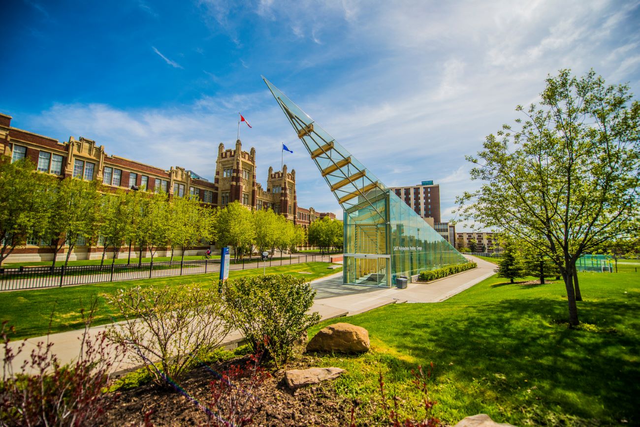 A modern glass pavilion with a triangular design set against a backdrop of historical buildings and greenery.