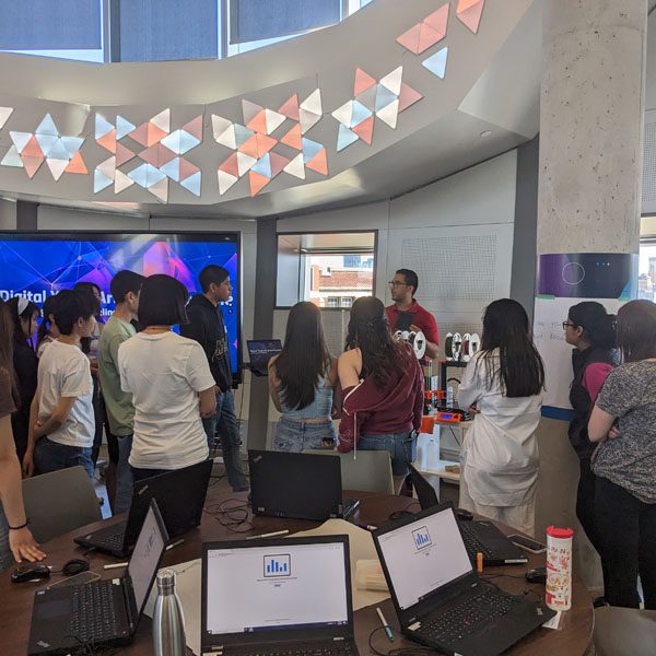 A group of students stands around an instructor giving a presentation in a modern classroom, with laptops open on tables and a large digital display screen in the background.