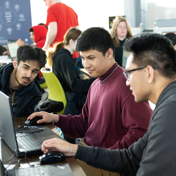 Three high school students collaborate on laptops in a classroom setting, discussing their work while other students study in the background.