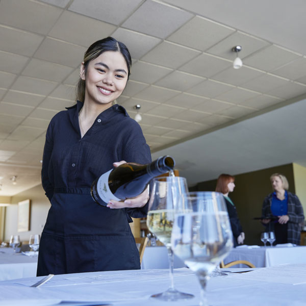 A smiling restaurant server wearing a black uniform pours white wine into a glass at a neatly set dining table, with other staff members visible in the background.