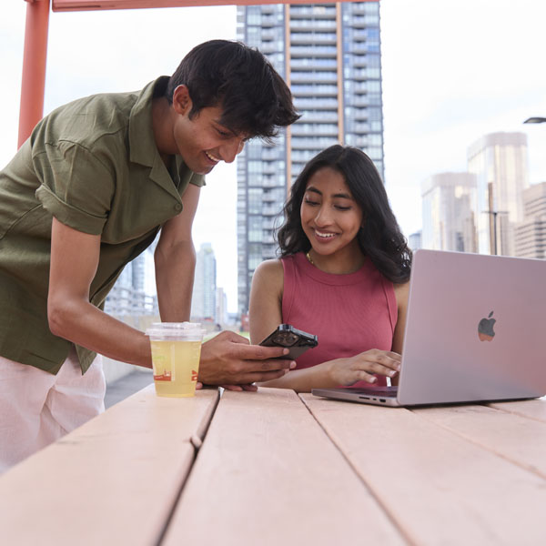 A young man and woman collaborate at a table outdoors, looking at a smartphone while a laptop sits open in front of the woman.