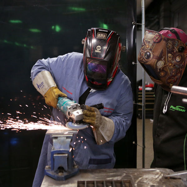 Two students wearing protective gear work together on a grinding task, with sparks flying from the equipment being used. 