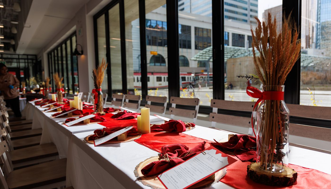 Long dining table set with red runners, menus, candles, and wheat centerpieces in a bright room with large windows.