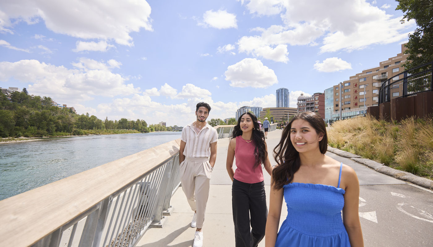 Three young adults walking along a riverside pathway on a sunny day, with buildings and trees in the background.