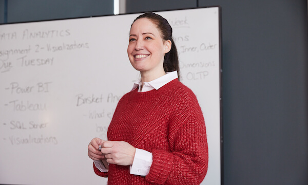 Smiling woman in a red sweater standing in front of a whiteboard with notes related to data analytics and visualization tools.