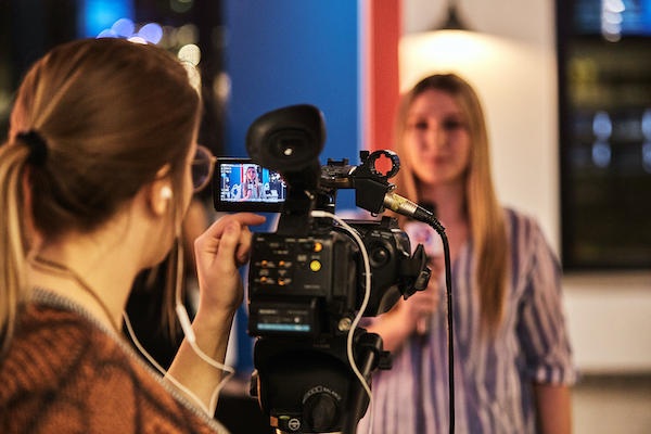 A reporter speaks into a microphone. A camera and cameraperson is seen in the foreground filming her.