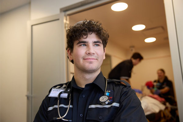 A young male paramedic wearing a uniform and stethoscope looks confidently while assisting in a medical setting.