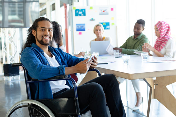 A smiling man in a wheelchair holds a phone while participating in a meeting with a diverse group of people.