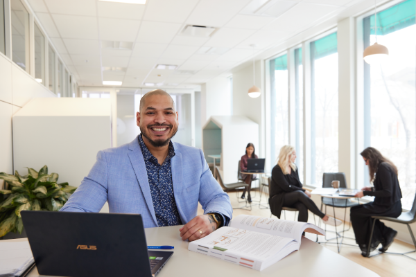 A smiling professional man in a blue blazer sits at a desk with a laptop and open book, while colleagues work in a modern office setting.