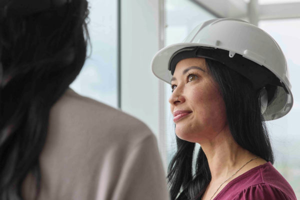 Woman wearing a white hard hat listens and speaks with a colleague in a bright office setting.