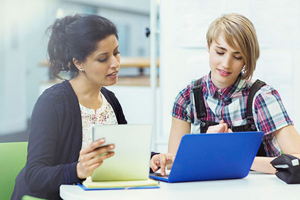 Two women have a discussion while looking at information on a laptop and tablet.