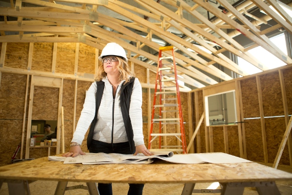 A woman in a hard hat stands at a table with construction plans in a framed building under construction.
