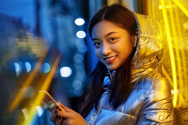 A young woman with long, black hair smiles at the camera while holding her phone. 