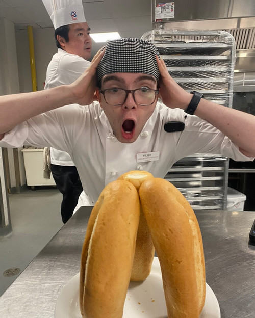 A surprised chef with glasses reacts dramatically to a stack of bread rolls.