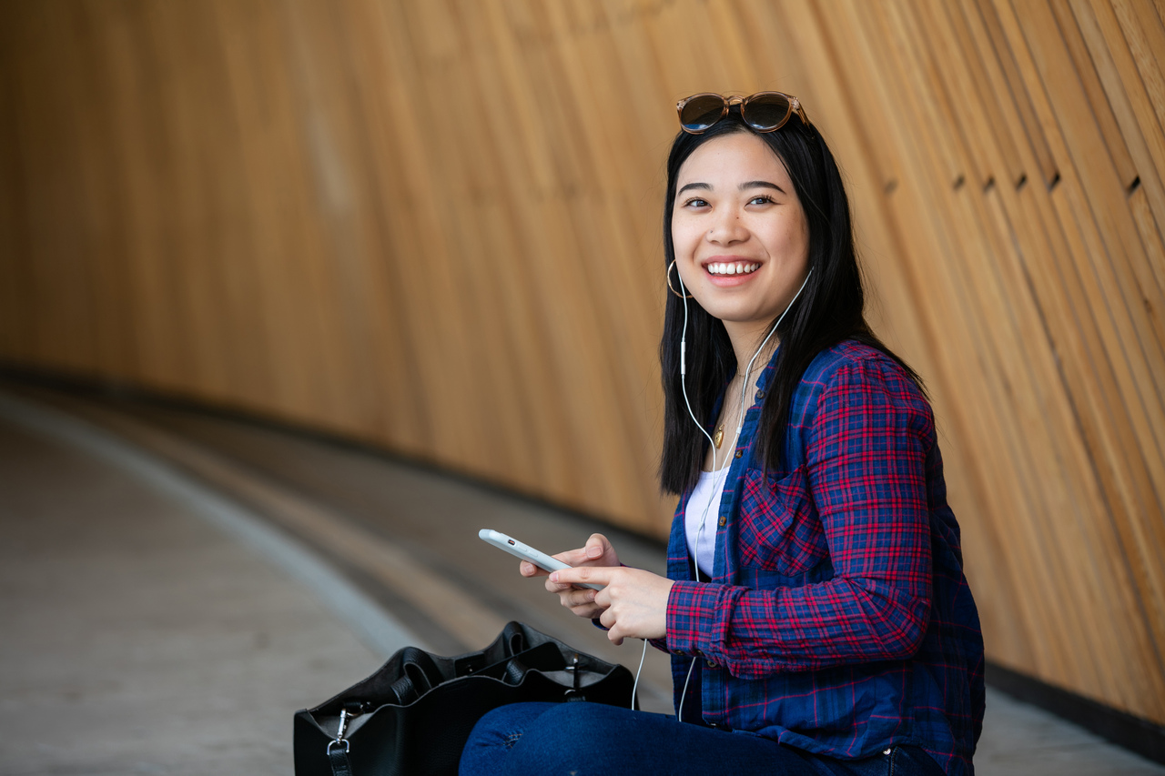 A young woman sitting against a wooden wall, smiling while using her smartphone and wearing headphones.