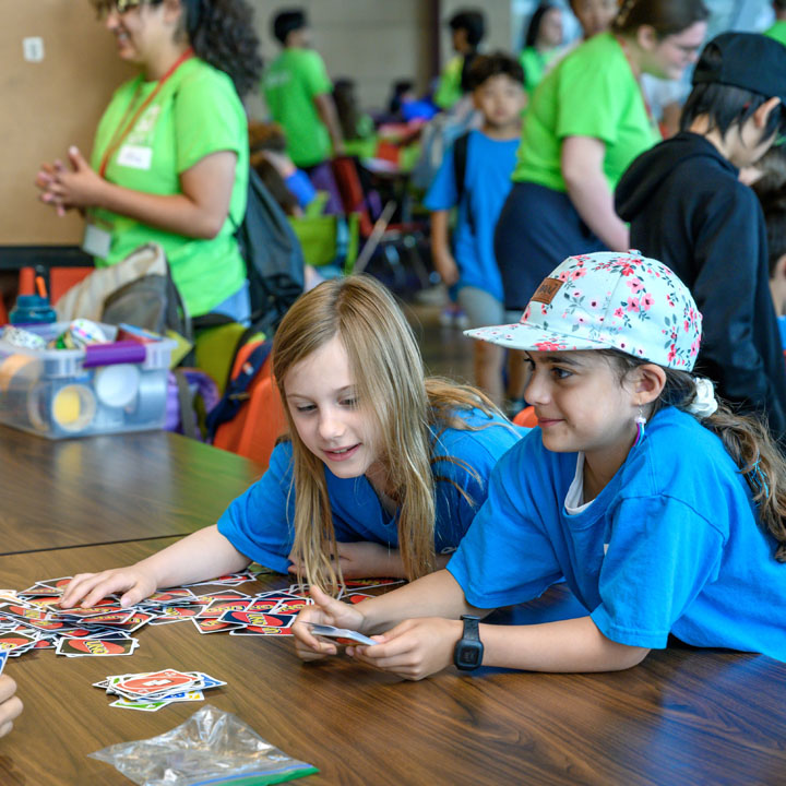 Two children wearing blue shirts play a card game at a table during a summer camp, while other kids and counselors in green shirts interact in the background.