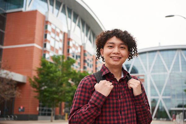 Person wearing a red plaid shirt and backpack standing outdoors on a modern campus, with glass-and-brick buildings in the background.