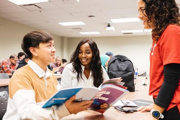Person wearing a red plaid shirt and backpack standing outdoors on a modern campus, with glass-and-brick buildings in the background.