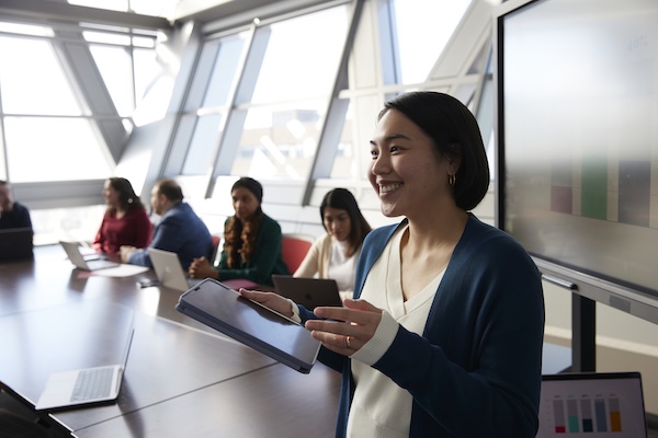 Smiling presenter holds a tablet while leading a meeting in a modern conference room with colleagues working on laptops in the background.
