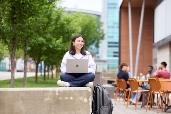 A student sits cross-legged outside on campus during the summertime, holding a laptop, smiling.