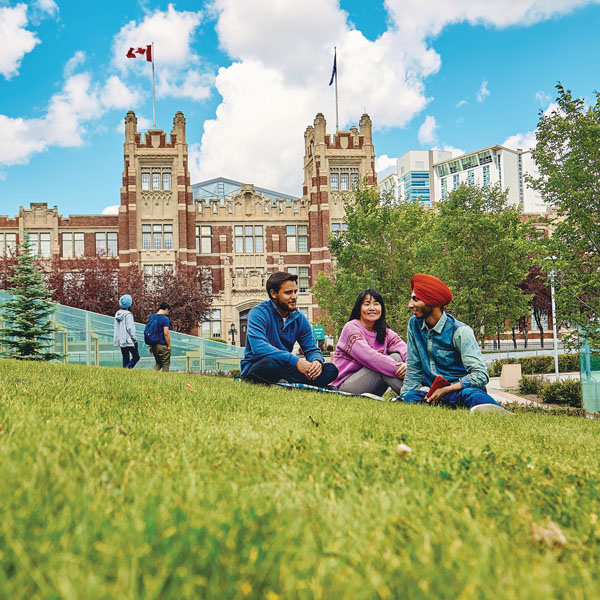 Three diverse students sitting on a grassy area in front of a historic building, enjoying a conversation on a sunny day.