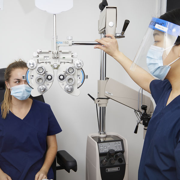 A healthcare professional performs an eye exam using a phoropter while a patient sits in a chair wearing a mask.