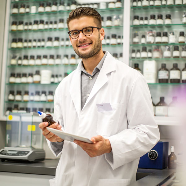 A smiling pharmacist in a lab coat holding a bottle and a clipboard, surrounded by shelves of pharmaceutical containers.