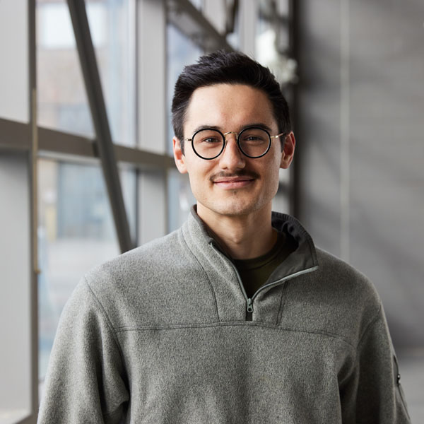 A young man with glasses and a mustache, wearing a gray fleece jacket, stands smiling in front of large windows.