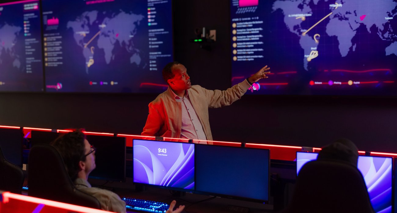 A man presents data on a digital map display in a high-tech command center, while two individuals attentively watch from their workstations.