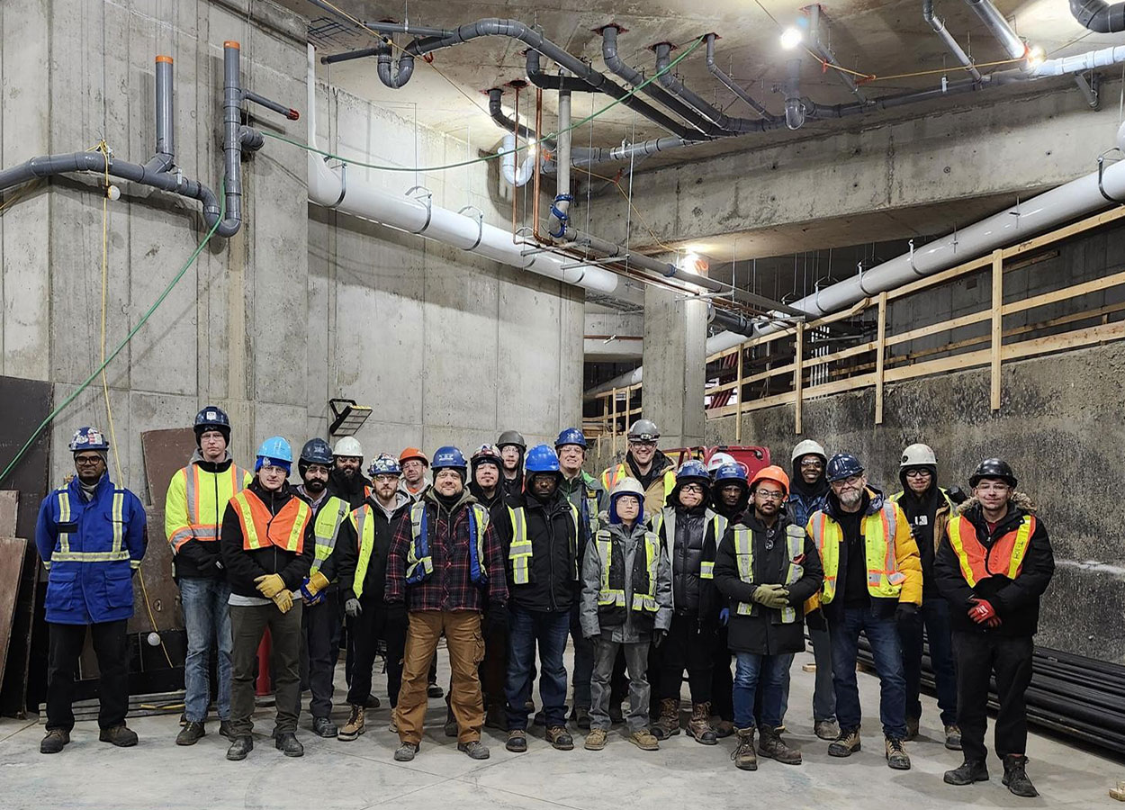 A group of fourth-year plumbing students and instructors at the Taylor Family Campus Centre construction site on Dec. 12, 2025. The group is standing in front of a concrete wall. 