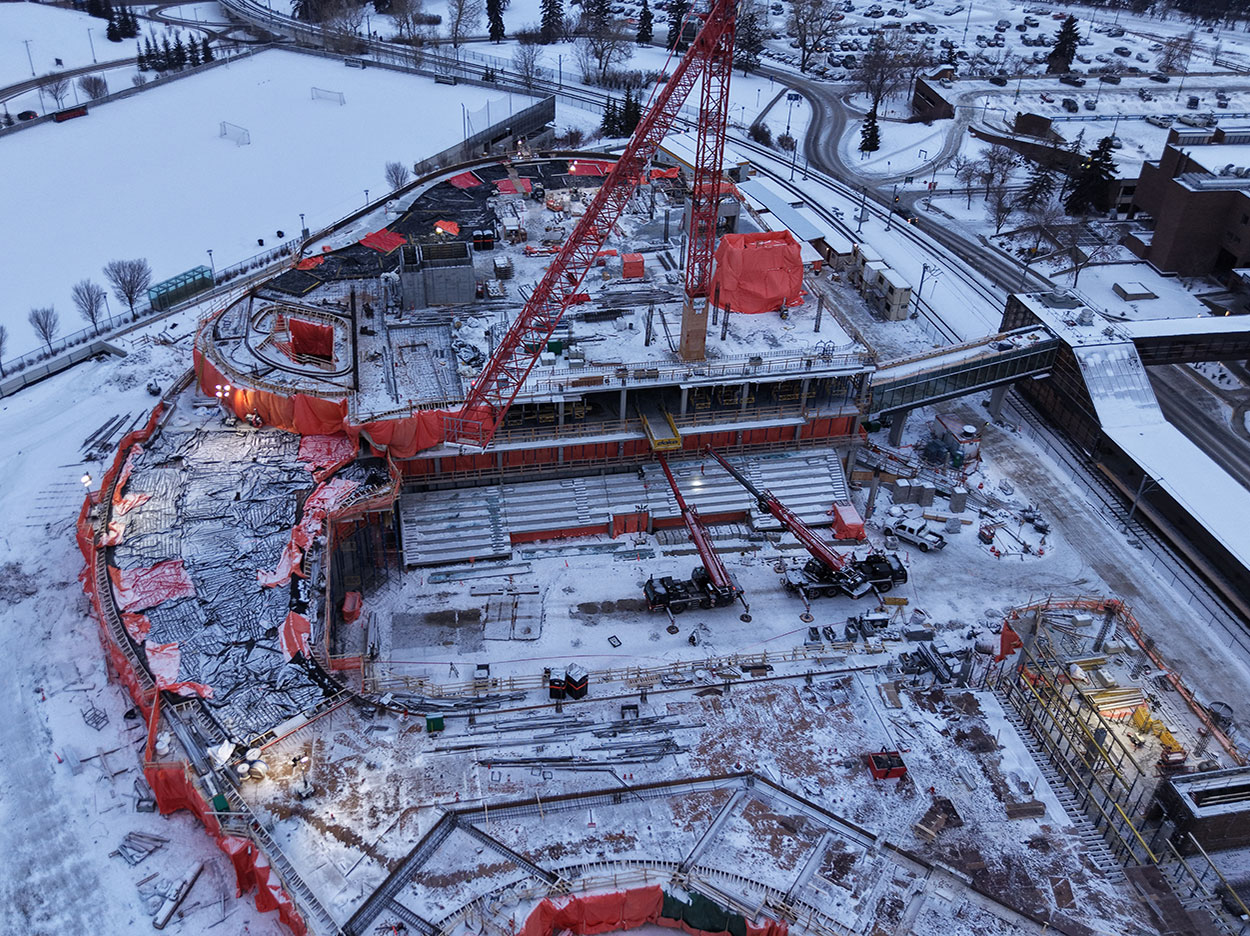 Aerial view of the Taylor Family Campus Centre construction site on Dec. 13, 2025, showing snow covering the ground, a large red crane, concrete superstructure, orange tarps and equipment around the building, in progress.