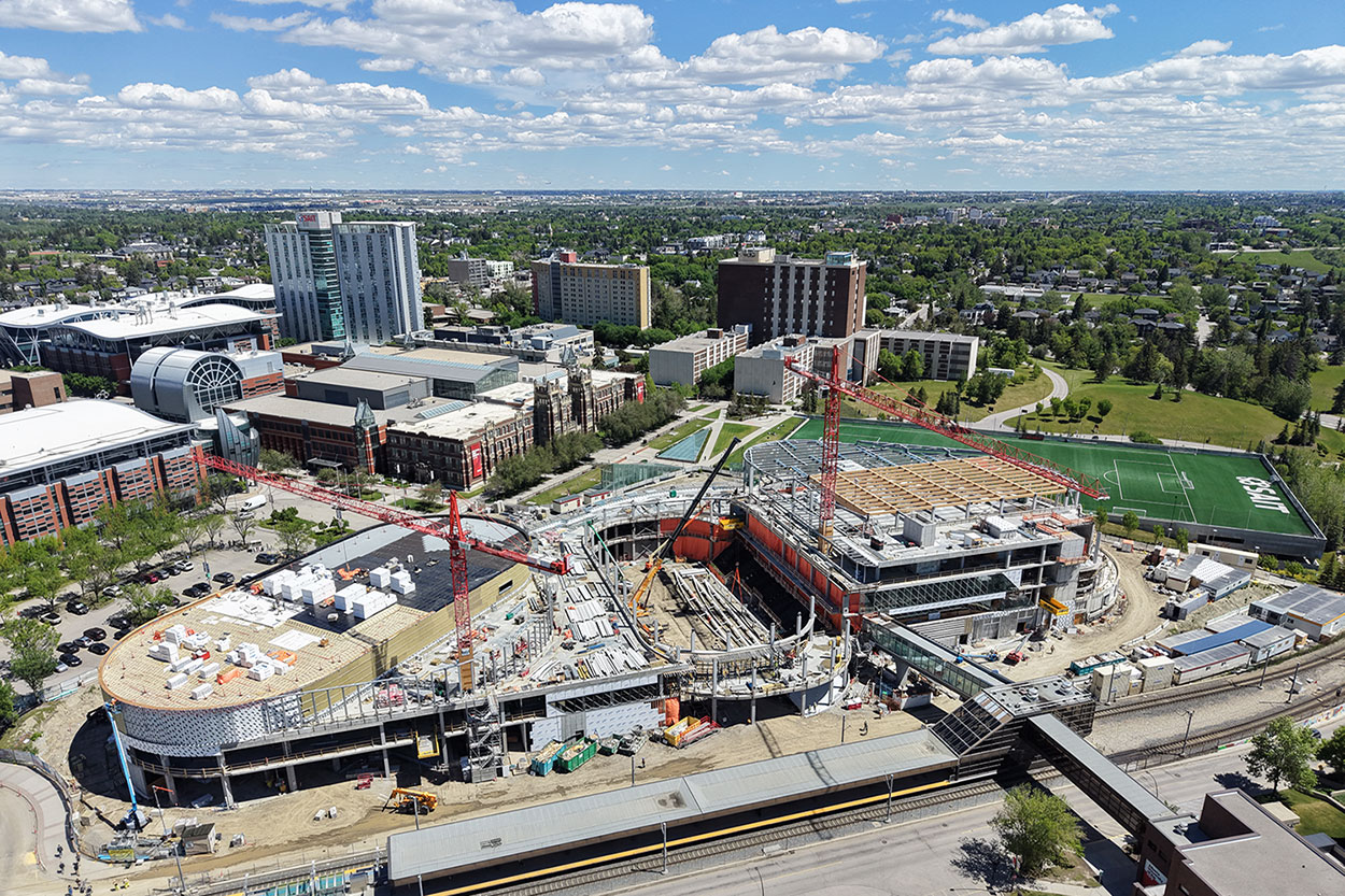 Aerial view of the Taylor Family Campus Centre construction site on June 8, 2026, showing large timber beams being installed on level three of the building, two large red cranes, concrete and steel superstructure, orange tarps and equipment around the building, in progress. The Cohos Commons soccer field is in the background, along with Heritage Hall and other campus buildings.