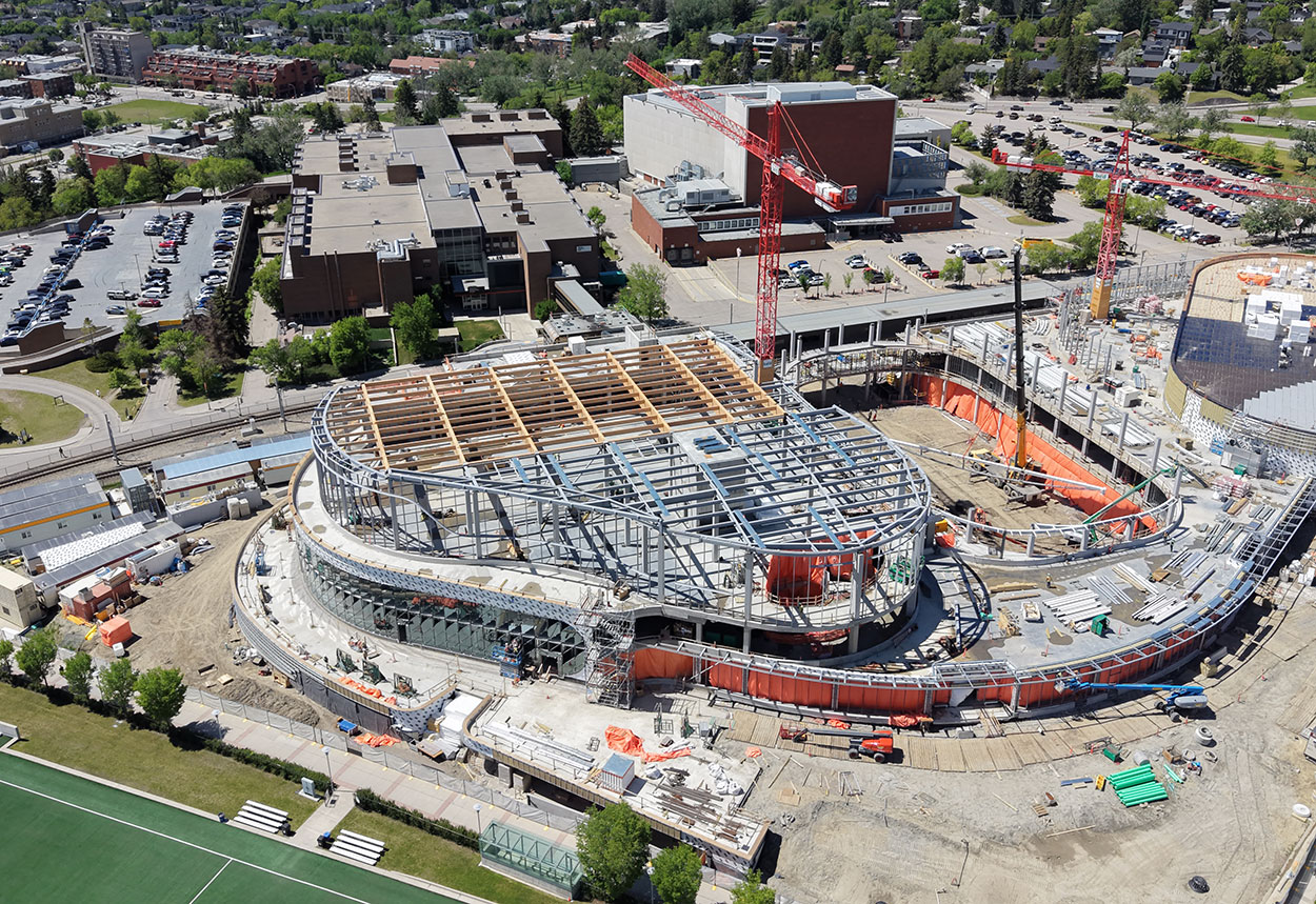 Aerial view of the Taylor Family Campus Centre construction site on June 8, 2026, showing large timber beams being installed on level three of the building, two large red cranes, concrete and steel superstructure, orange tarps and equipment around the building, in progress. The Southern Alberta Jubilee Auditorium and the Alberta University for the Arts buildings are in the background, along with the CTrain tracks.