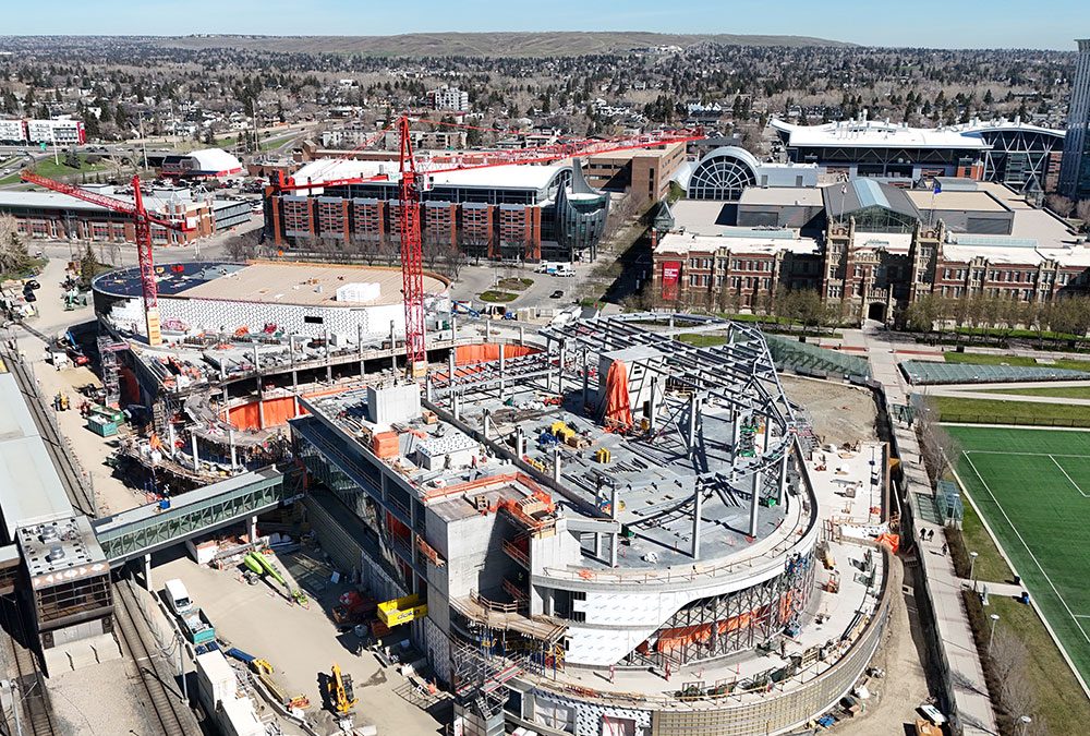 Aerial view of the Taylor Family Campus Centre construction site on May 3, 2026, showing two large red cranes, concrete and steel superstructure, orange tarps and equipment around the building, in progress.