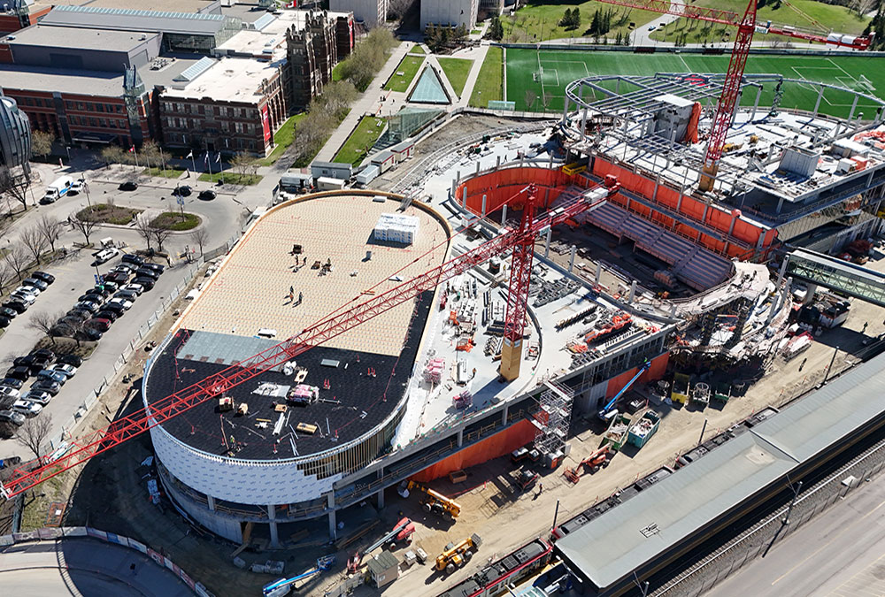 Aerial view of the Taylor Family Campus Centre construction site on May 3, 2026, showing two large red cranes, the roof of the gym under construction, and steel and concrete superstructure, and workers around the building, in progress.