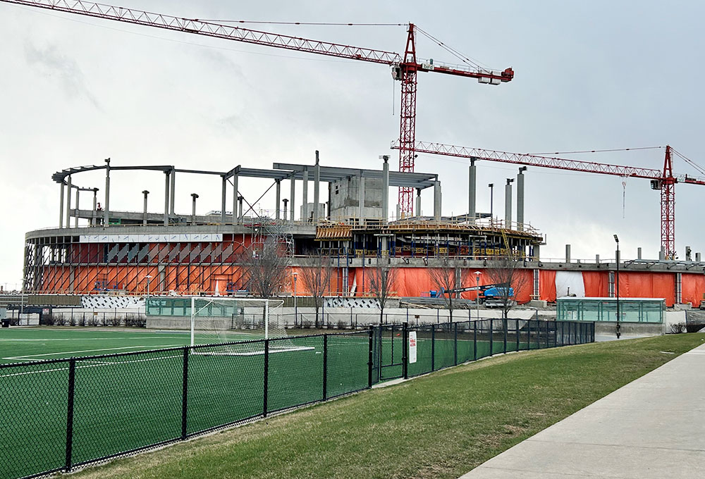 Taylor Family Campus Centre under construction April 23, 2026, showing two large red cranes, concrete superstructure, orange tarps and equipment around the building, in progress.