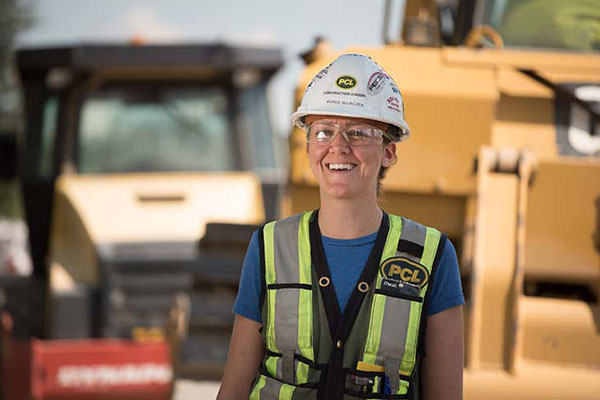 A woman wearing a hardhat, safety glasses and safety vest smiles at the camera on a construction site.