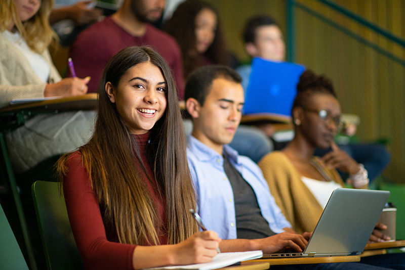 A diverse group of students seated in a large lecture hall, smiling and engaging with the instructor or the topic, surrounded by tiered rows of seats and ambient campus lighting.