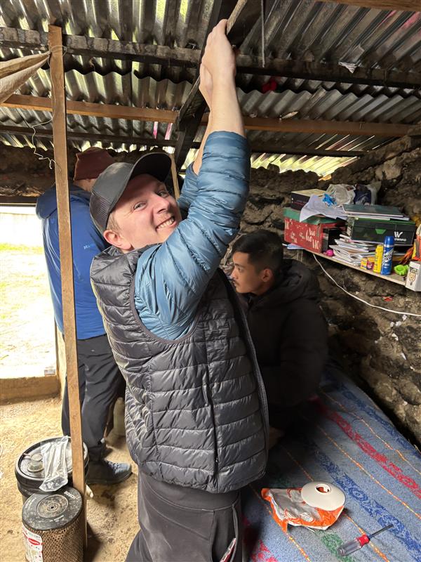 A man in a blue jacket and black hat is tightening a strap while another person sits nearby in a rustic setting.