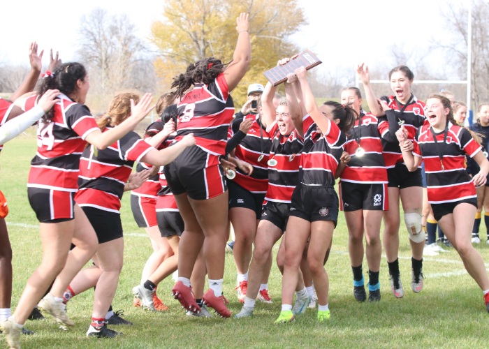 Women’s rugby team in red-and-black striped jerseys celebrates on a grassy field, with one player holding a trophy overhead.