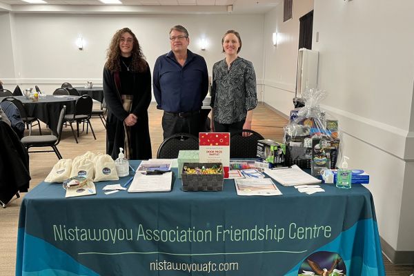 Three individuals standing in front of a table with informational material at the Nistawoyou Association Friendship Centre.