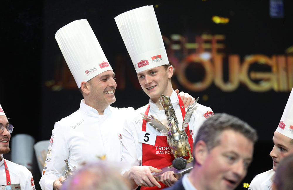 Chef in a professional kitcåTwo chefs celebrating their victory at a cooking competition, wearing tall white hats and holding trophies, with other participants in the background.hen giving a cooking demonstration to a small group.