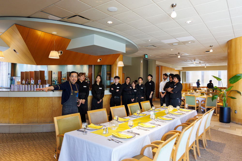 A restaurant staff member gestures to a beautifully set dining table, while a team of waitstaff stands ready in the background