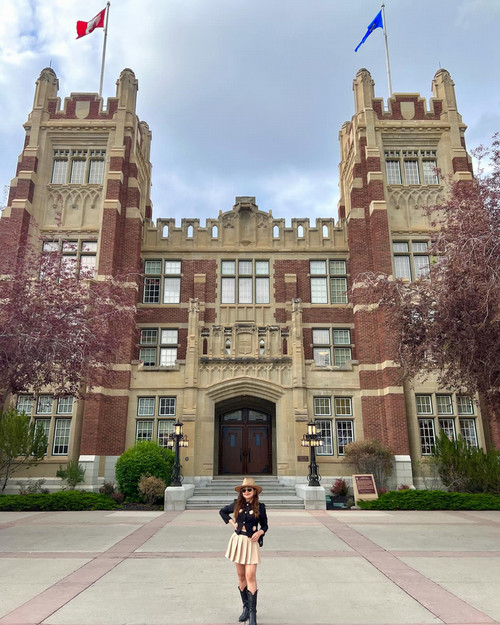 A student in a black jacket and striped skirt stands in front of Heritage Hall, a historic brick building with two towers and the Canadian and Albertan flags flying.