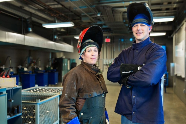 Two welders in protective gear stand side by side in a workshop, helmets raised and smiling at the camera.