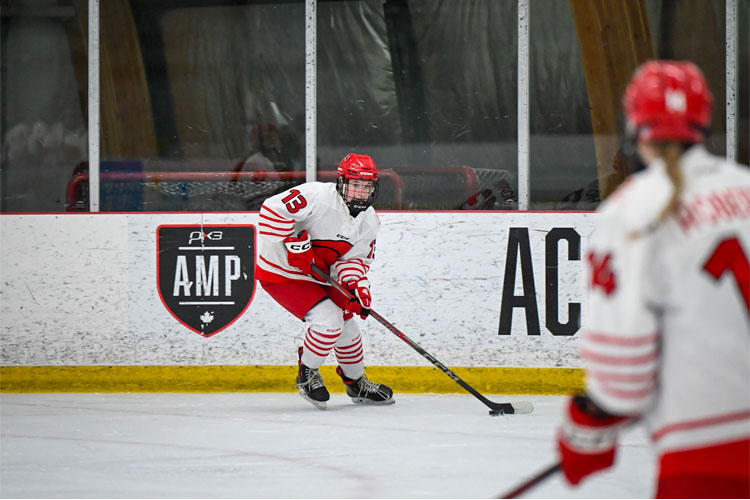 Trojans women's hockey player in a red helmet and white-and-red uniform skates with the puck along the boards during a game, with a blurred teammate in the foreground.