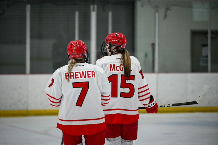 Two Trojans women's ice hockey players in white-and-red jerseys, numbers 7 and 15, stand on the rink with their backs to the camera.