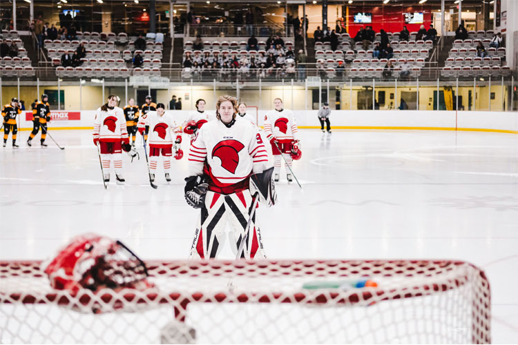 Trojans women's hockey goalie in white-and-red gear stands on the rink facing the camera, with teammates lined up behind and empty arena seats in the background.