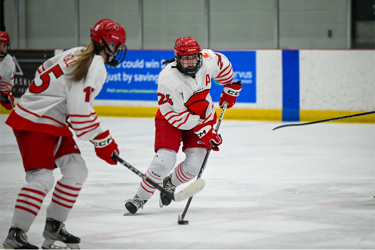 Two Trojans women's ice hockey players in white-and-red uniforms skate on an indoor rink, with one controlling the puck on their stick.